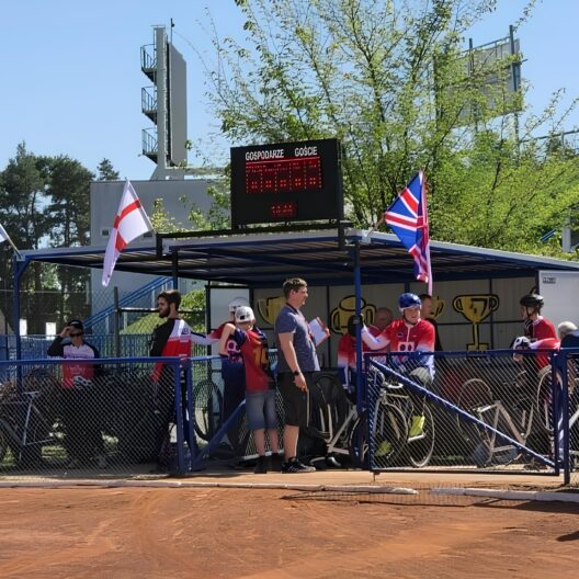 Group of Athletes in Red Uniforms Standing Behind Blue Fencing at an Outdoor Sports Event with a Digital Scoreboard and Flags in the Background 9 Września 2025 
