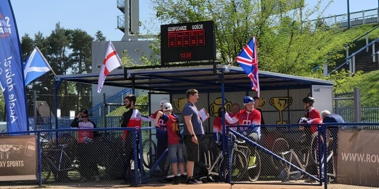 Group of Athletes in Red Uniforms Standing Behind Blue Fencing at an Outdoor Sports Event with a Digital Scoreboard and Flags in the Background 5 Maja 2026 