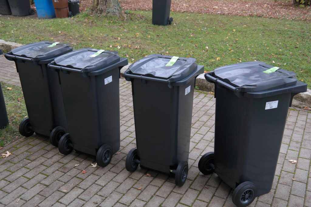 Four Black Wheeled Trash Bins with Closed Lids Lined Up on a Brick Path Beside a Grassy Area with Fallen Leaves in Autumn 9 Września 2025
