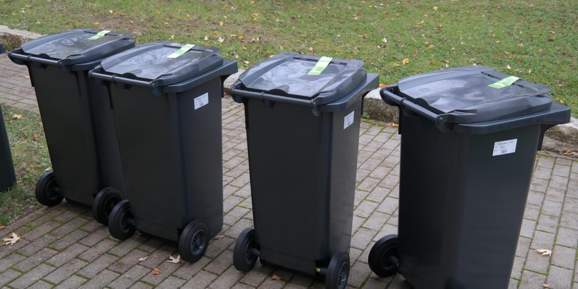 Four Black Wheeled Trash Bins with Closed Lids Lined Up on a Brick Path Beside a Grassy Area with Fallen Leaves in Autumn 15 Czerwca 2020