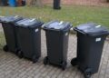 Four Black Wheeled Trash Bins with Closed Lids Lined Up on a Brick Path Beside a Grassy Area with Fallen Leaves in Autumn 15 Czerwca 2020