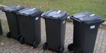 Four Black Wheeled Trash Bins with Closed Lids Lined Up on a Brick Path Beside a Grassy Area with Fallen Leaves in Autumn 15 Czerwca 2020