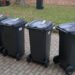 Four Black Wheeled Trash Bins with Closed Lids Lined Up on a Brick Path Beside a Grassy Area with Fallen Leaves in Autumn 15 Czerwca 2020