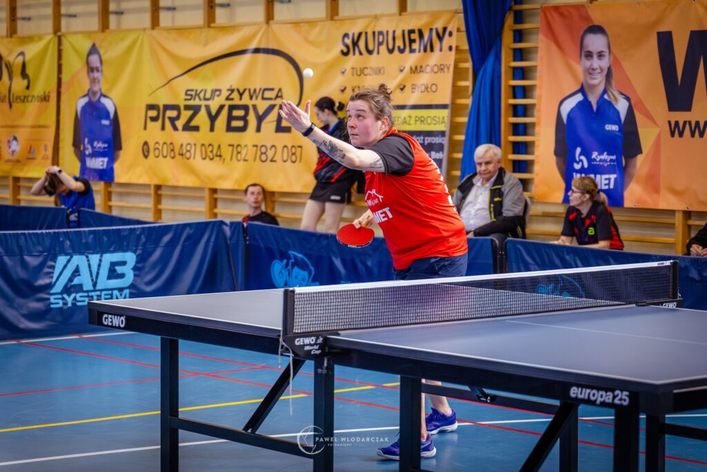 Female Table Tennis Player in a Red Shirt Reaching to Return the Ball During a Match in an Indoor Gym with Banners in the Background 9 Września 2025 
