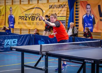 Female Table Tennis Player in a Red Shirt Reaching to Return the Ball During a Match in an Indoor Gym with Banners in the Background 15 Września 2023 