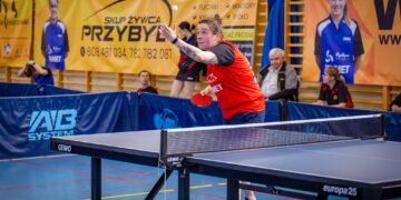 Female Table Tennis Player in a Red Shirt Reaching to Return the Ball During a Match in an Indoor Gym with Banners in the Background 15 Czerwca 2020
