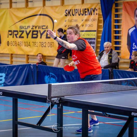 Female Table Tennis Player in a Red Shirt Reaching to Return the Ball During a Match in an Indoor Gym with Banners in the Background 9 Września 2025 