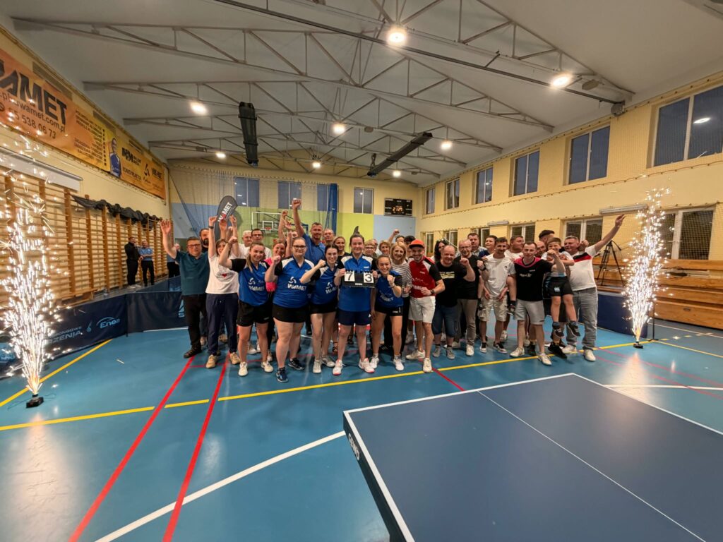 Group of Athletes Posing for a Celebration Photo in a Gymnasium Many Raising Their Arms and Cheering with Banners on the Wall Behind Them and Sparklers at the Sides 9 Września 2025