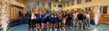 Group of Athletes Posing for a Celebration Photo in a Gymnasium Many Raising Their Arms and Cheering with Banners on the Wall Behind Them and Sparklers at the Sides 25 Maja 2026