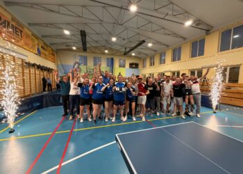 Group of Athletes Posing for a Celebration Photo in a Gymnasium Many Raising Their Arms and Cheering with Banners on the Wall Behind Them and Sparklers at the Sides 13 Sierpnia 2023