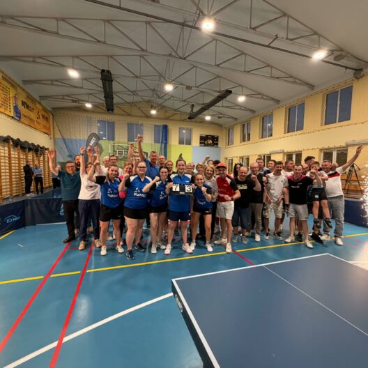 Group of Athletes Posing for a Celebration Photo in a Gymnasium Many Raising Their Arms and Cheering with Banners on the Wall Behind Them and Sparklers at the Sides 9 Września 2025