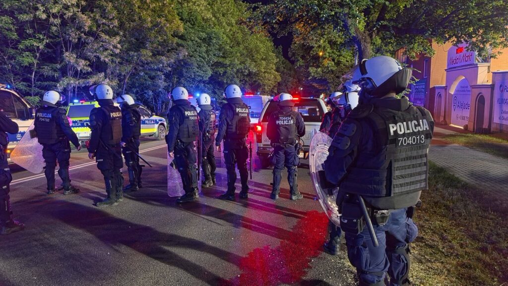 Line of Riot Police in Body Armor and Helmets with Shields Standing on a City Street at Night Blue and Red Flashing Patrol Lights in the Background 9 Września 2025