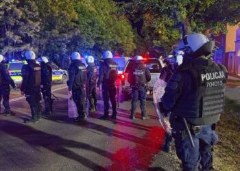 Line of Riot Police in Body Armor and Helmets with Shields Standing on a City Street at Night Blue and Red Flashing Patrol Lights in the Background 10 Lipca 2019