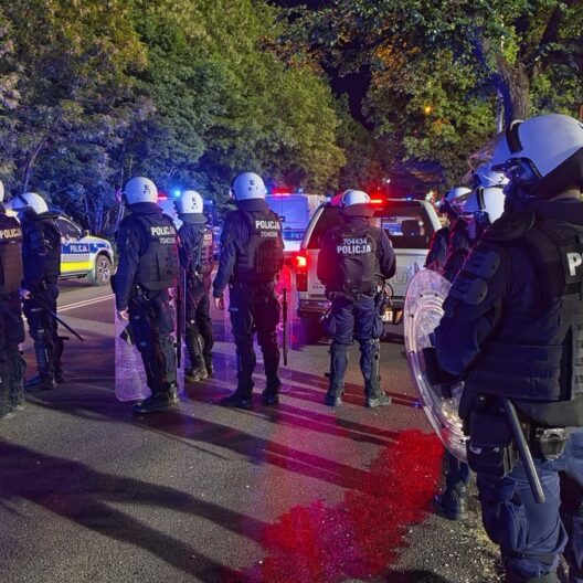Line of Riot Police in Body Armor and Helmets with Shields Standing on a City Street at Night Blue and Red Flashing Patrol Lights in the Background 9 Września 2025