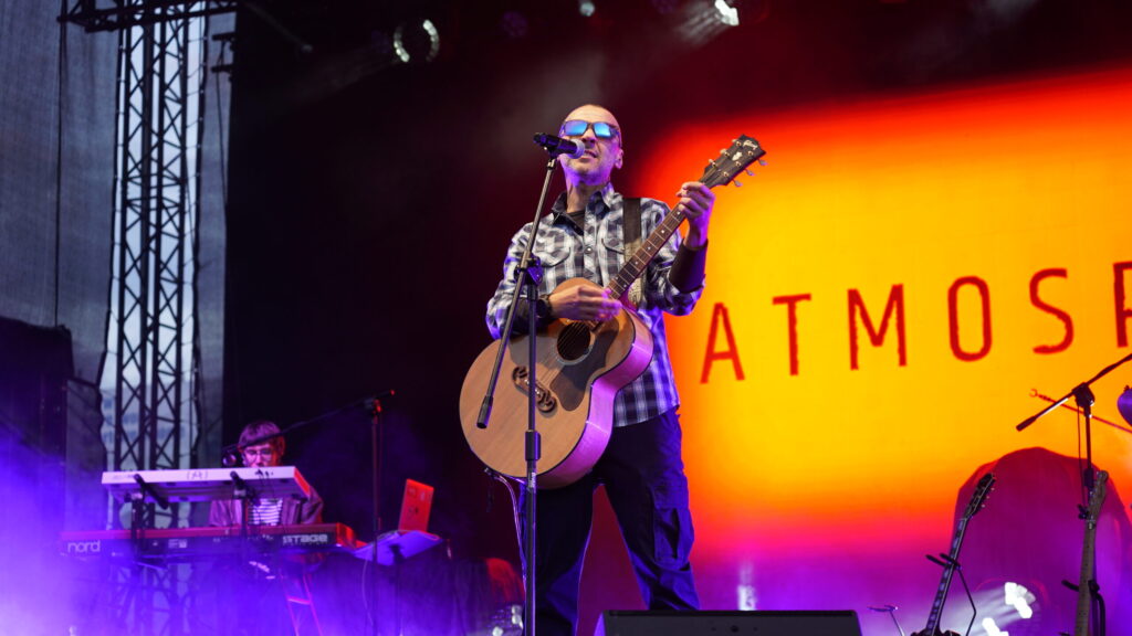 Male Musician Wearing Sunglasses and a Plaid Shirt Sings into a Microphone While Playing an Acoustic Guitar on Stage with Purple Fog and an Orange Backdrop Reading Atmos 9 Września 2025