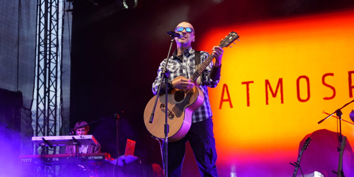 Male Musician Wearing Sunglasses and a Plaid Shirt Sings into a Microphone While Playing an Acoustic Guitar on Stage with Purple Fog and an Orange Backdrop Reading Atmos 16 Czerwca 2020