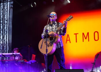 Male Musician Wearing Sunglasses and a Plaid Shirt Sings into a Microphone While Playing an Acoustic Guitar on Stage with Purple Fog and an Orange Backdrop Reading Atmos 16 Września 2025