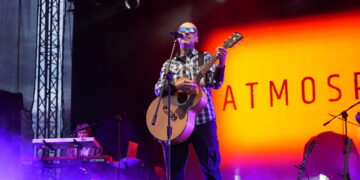 Male Musician Wearing Sunglasses and a Plaid Shirt Sings into a Microphone While Playing an Acoustic Guitar on Stage with Purple Fog and an Orange Backdrop Reading Atmos 14 Czerwca 2020
