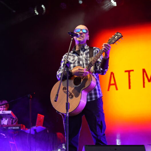 Male Musician Wearing Sunglasses and a Plaid Shirt Sings into a Microphone While Playing an Acoustic Guitar on Stage with Purple Fog and an Orange Backdrop Reading Atmos 9 Września 2025