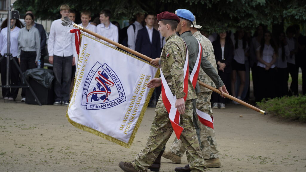 Soldiers in Camouflage March While Carrying a Large Ceremonial Flag with Red and white Ribbons at an Outdoor Event a Crowd Stands in the Background 9 Września 2025