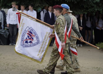 Soldiers in Camouflage March While Carrying a Large Ceremonial Flag with Red and white Ribbons at an Outdoor Event a Crowd Stands in the Background 16 Września 2023