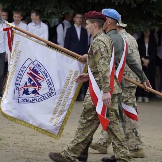 Soldiers in Camouflage March While Carrying a Large Ceremonial Flag with Red and white Ribbons at an Outdoor Event a Crowd Stands in the Background 9 Września 2025