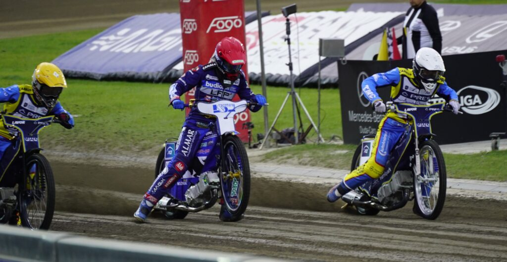 Three Speedway Riders in Blue and Yellow Suits Race on a Muddy Track Kicking Up Dirt As They Round a Bend with Banners in the Background 9 Września 2025