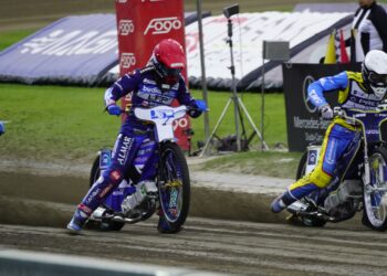 Three Speedway Riders in Blue and Yellow Suits Race on a Muddy Track Kicking Up Dirt As They Round a Bend with Banners in the Background 13 Sierpnia 2023