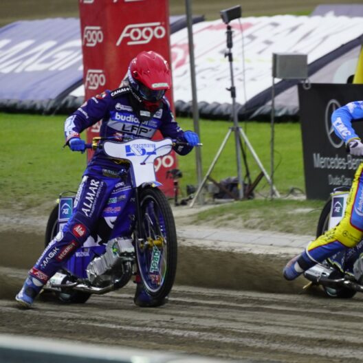 Three Speedway Riders in Blue and Yellow Suits Race on a Muddy Track Kicking Up Dirt As They Round a Bend with Banners in the Background 9 Września 2025