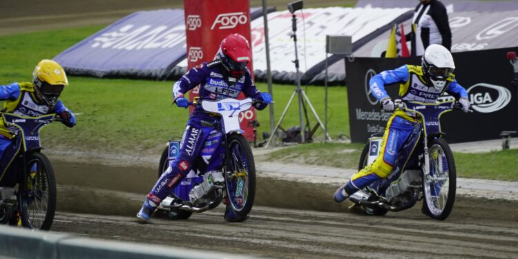 Three Speedway Riders in Blue and Yellow Suits Race on a Muddy Track Kicking Up Dirt As They Round a Bend with Banners in the Background 13 Czerwca 2026